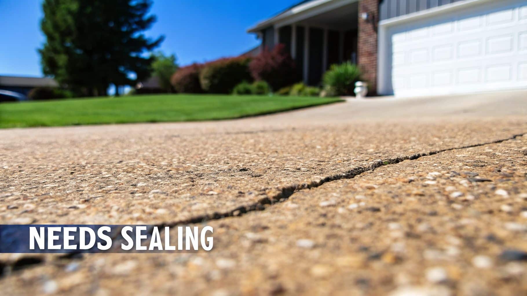 Close-up of a cracked concrete driveway needing sealing, with a house and green lawn.
