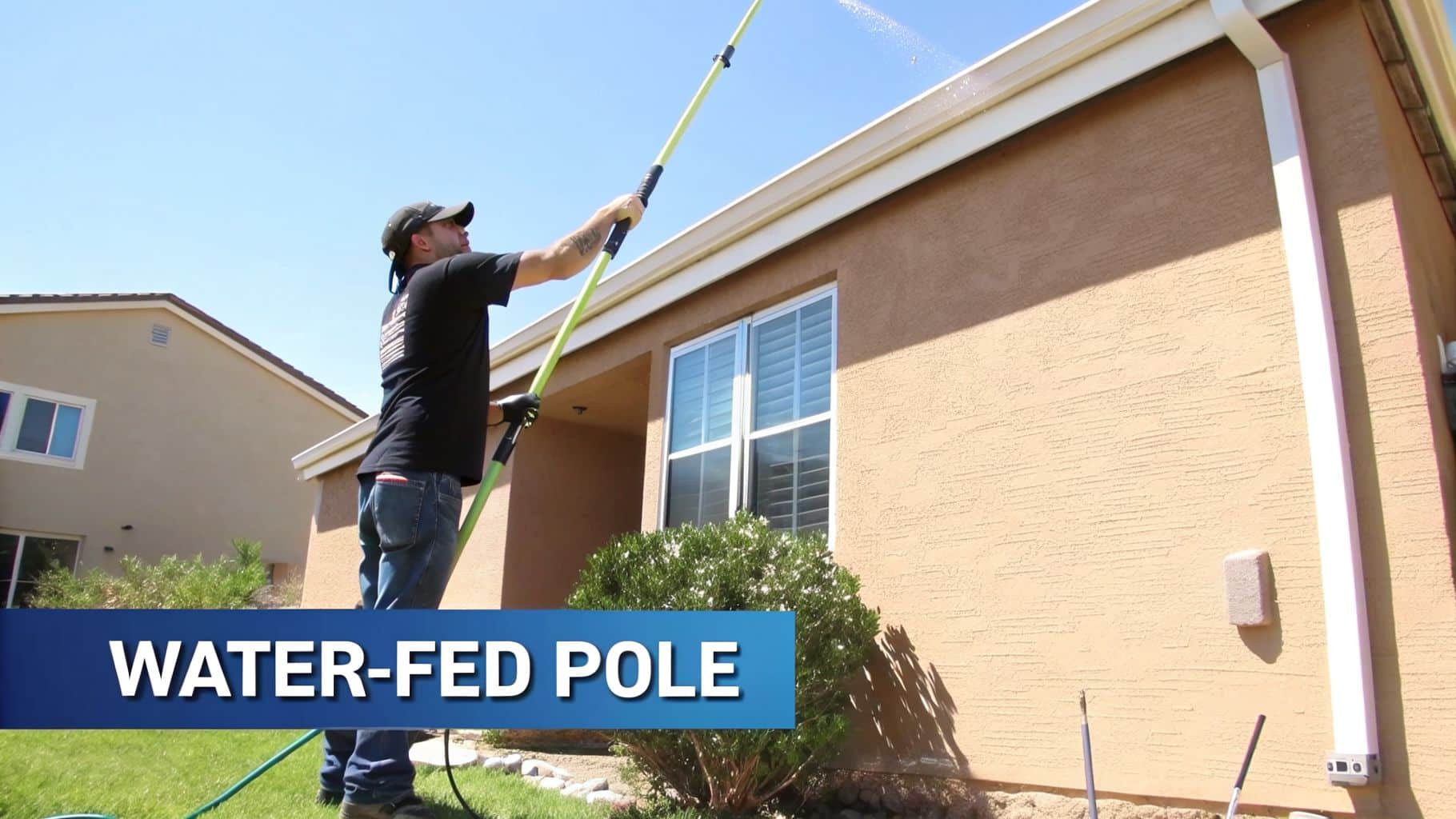 A person uses a long, water-fed pole to spray and clean the high gutters of a house.