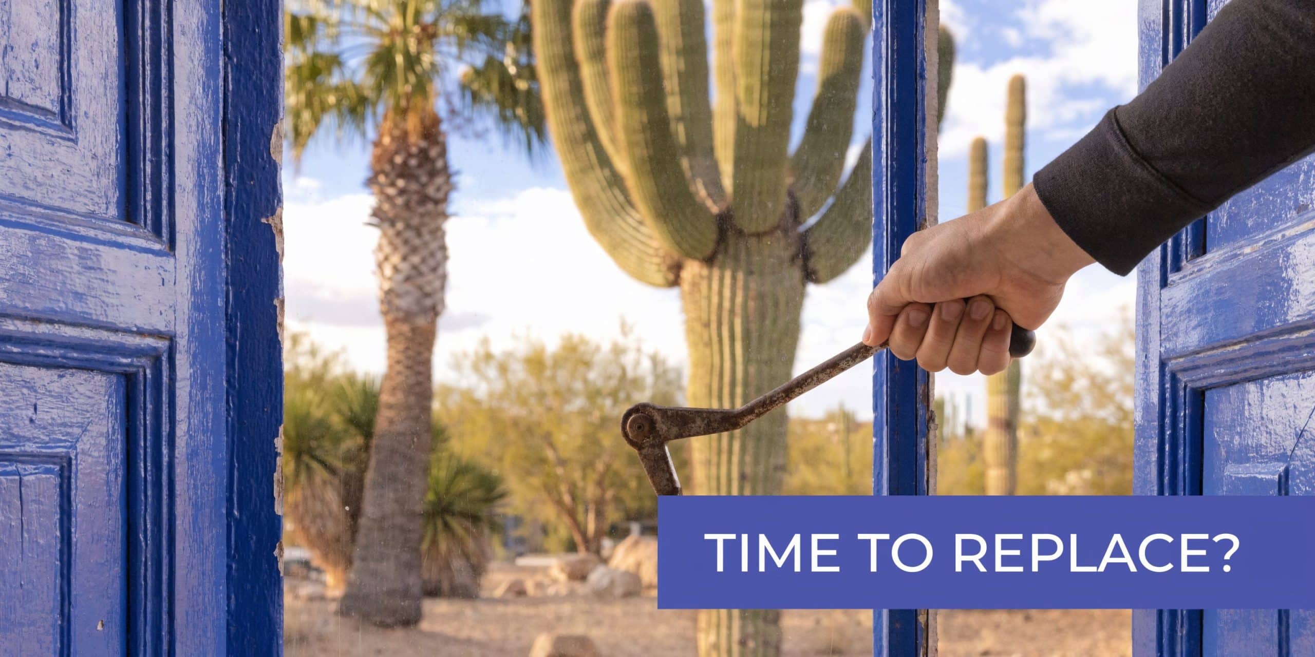 A hand holds a latch on an old, weathered blue window frame with a desert cactus landscape background.