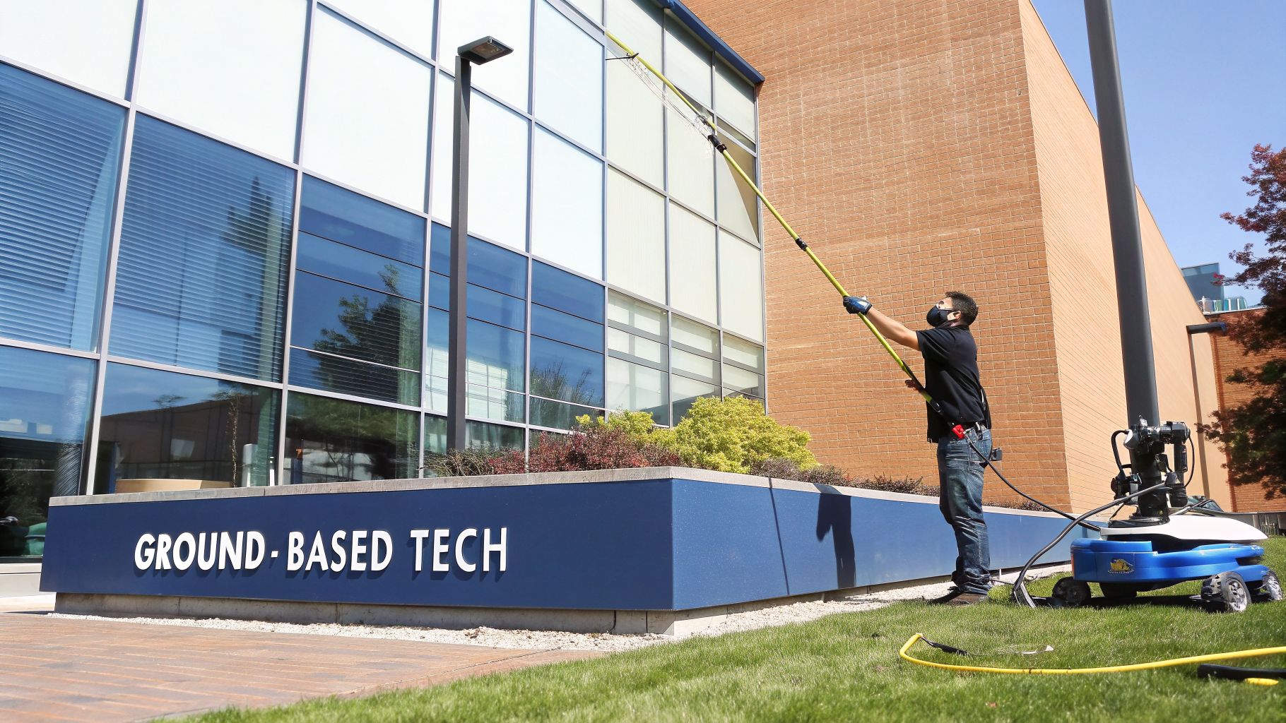 A worker in a mask and gloves cleans modern building windows with a long pole and ground-based tech.